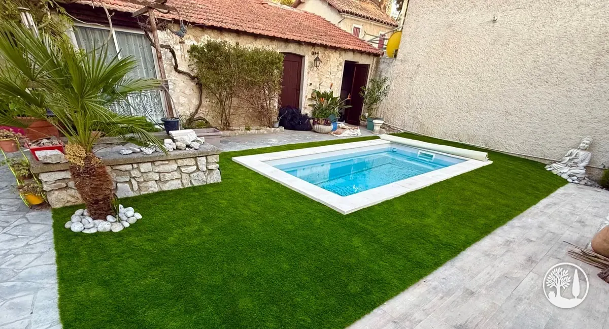 Gazon synthétique posé autour d'une piscine, palmier et bordure de galets blancs